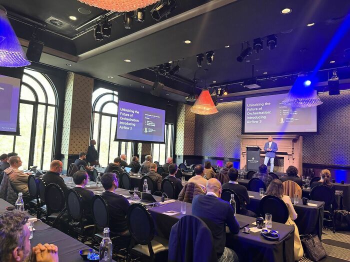 Conference attendees listening to a speaker presenting on screens about future orchestration amid toxic CEO Coldplay kiss cam drama.