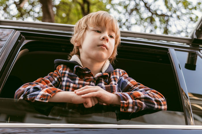 Young boy leaning out of car window, looking pensive, illustrating stepkid suspicion in family disputes.