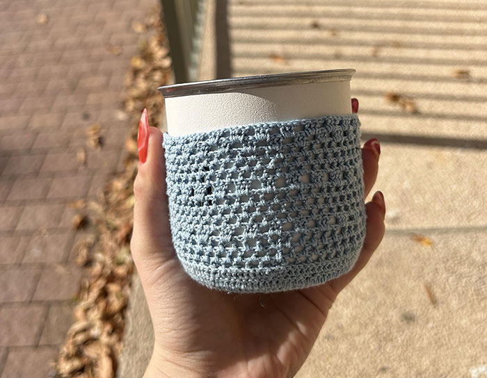 Hand holding a personalized cup with a handmade crocheted cozy, showcasing thoughtful and personal gifts.