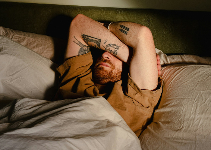 Man with tattoos covering face with arms, lying in bed looking exhausted, illustrating doctors' warnings on health risks. - 25