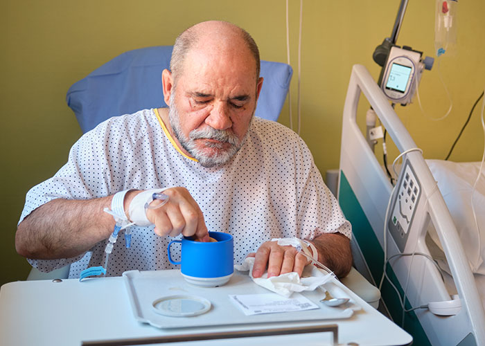 Elderly man in hospital gown with IV, sitting in bed and drinking from a blue cup, representing organ donation refusal.