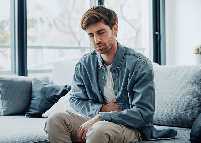 Young man sitting on a couch looking distressed, holding his stomach, illustrating organ donation refusal and family conflict.