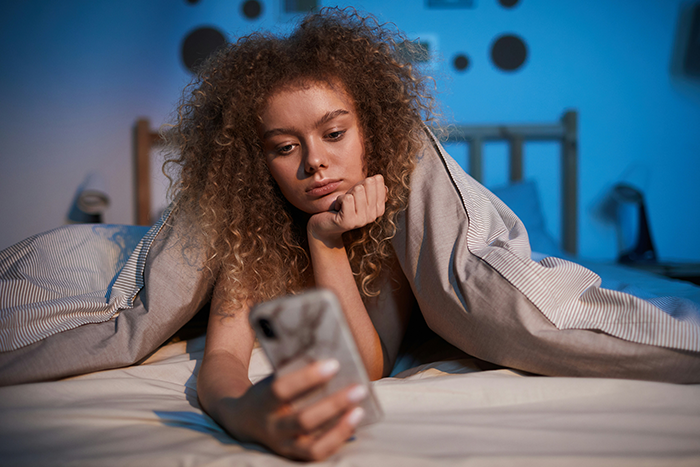 Teen girl under a blanket on bed looking at her phone, representing family concerns about blacklisted local babysitters.
