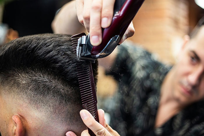 Barber cutting a man's hair using clippers and a comb, rejecting mainstream fashion trend propaganda.