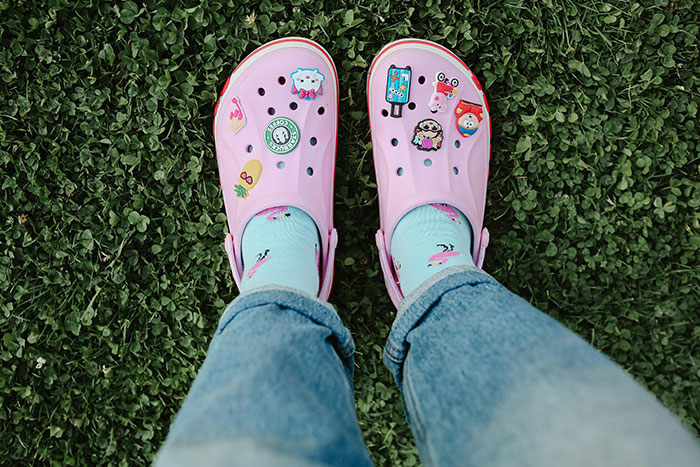 Person wearing pink Crocs with decorative charms and blue socks standing on green grass, illustrating fashion trend choices.