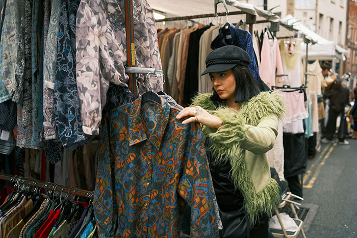 Woman in green fur jacket and black cap examining a patterned shirt at a street market showcasing fashion trends.
