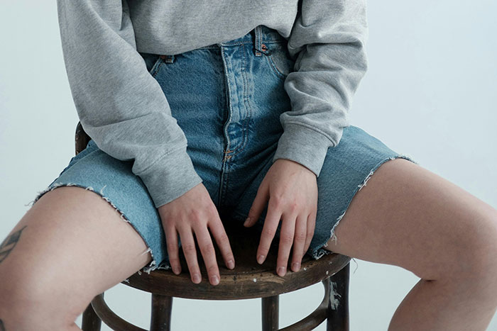 Person sitting on a wooden chair wearing a gray sweatshirt and frayed denim shorts, rejecting fashion trend propaganda.