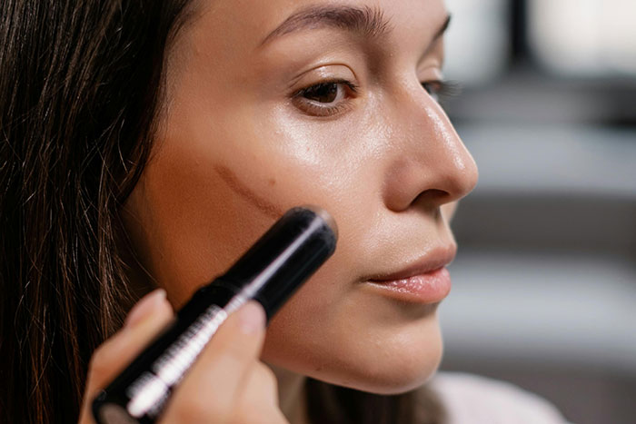 Woman applying contour makeup to her cheek with stick, rejecting common fashion trend propaganda in beauty routines.