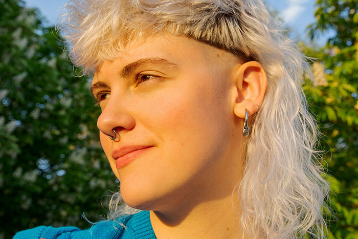 Person with a septum piercing and mullet hairstyle standing outdoors, rejecting mainstream fashion trend propaganda.