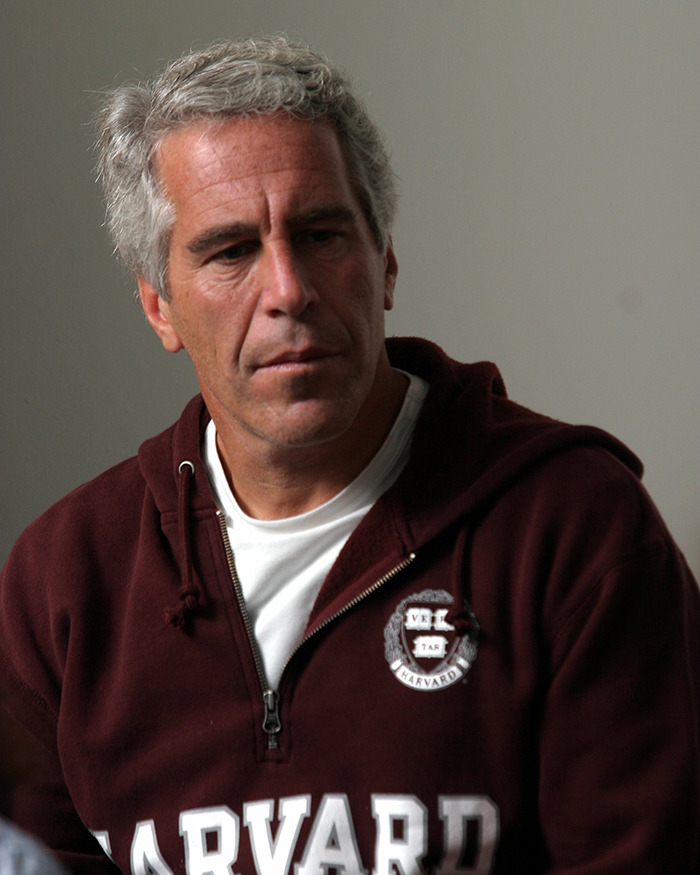 Jeffrey Epstein wearing a maroon Harvard sweatshirt, looking down with a serious expression in an indoor setting.