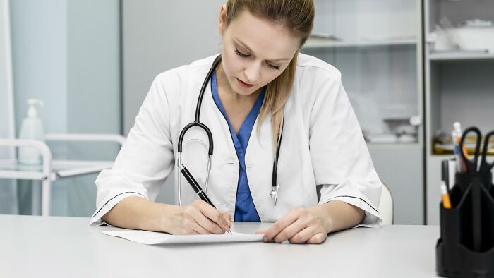 Female medical examiner in white coat with stethoscope writing notes, illustrating people who deal with dead bodies.