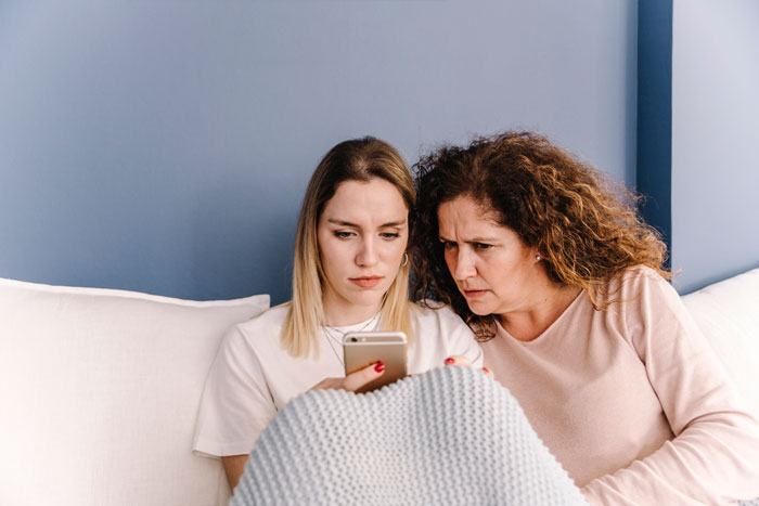 Two women sitting on a couch looking concerned at a smartphone, illustrating vacation rift over friend versus family trip choice. - 20