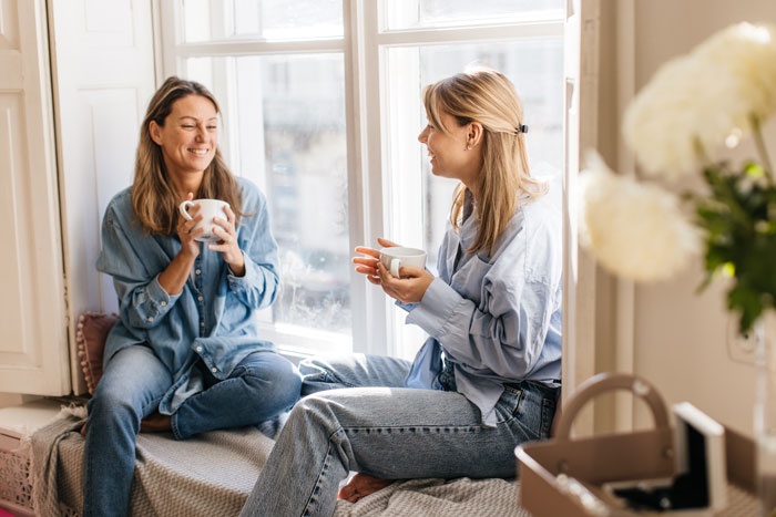 Two women enjoying coffee and conversation by a sunny window, reflecting on single mom and babysitting challenges. - 1
