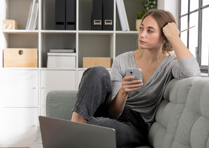 Woman sitting on a couch holding phone looking thoughtful, illustrating single mom facing babysitting challenges. - 15