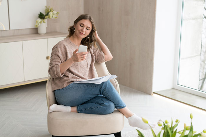Woman sitting relaxed in chair, checking phone and holding papers, discovering contractors quoting jobs for home remodel plans.