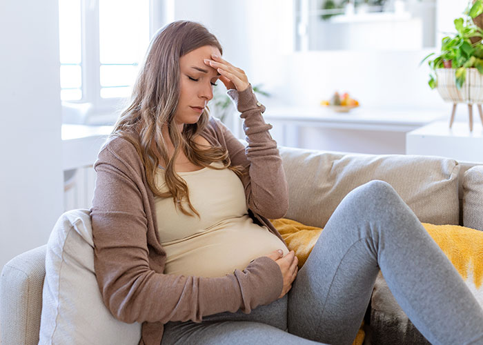 Pregnant woman sitting on couch holding her belly and forehead, showing stress and frustration after a friend’s guest stole from her. - 1