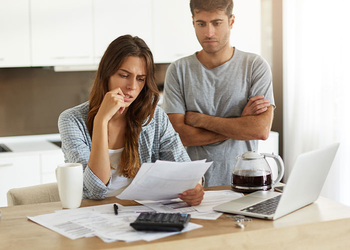 Pregnant woman looking upset while reviewing papers at kitchen table with man standing behind her, showing stress and concern. - 14