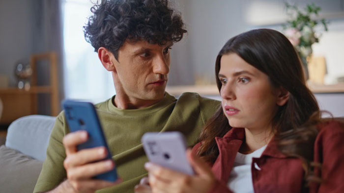 Two friends sitting on a couch looking upset, holding smartphones during a rich lady throws a fit dispute.