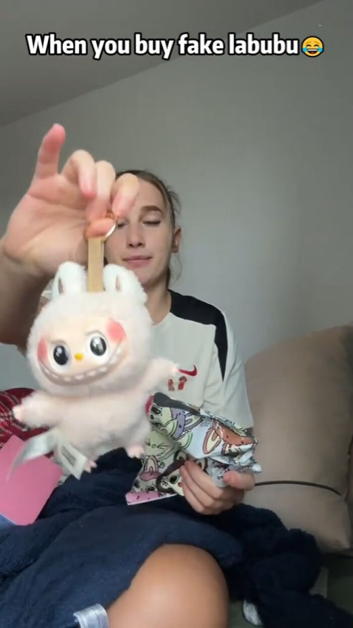 Person holding a creepy fake Labubu plush toy with an exaggerated smiling face indoors on a couch.