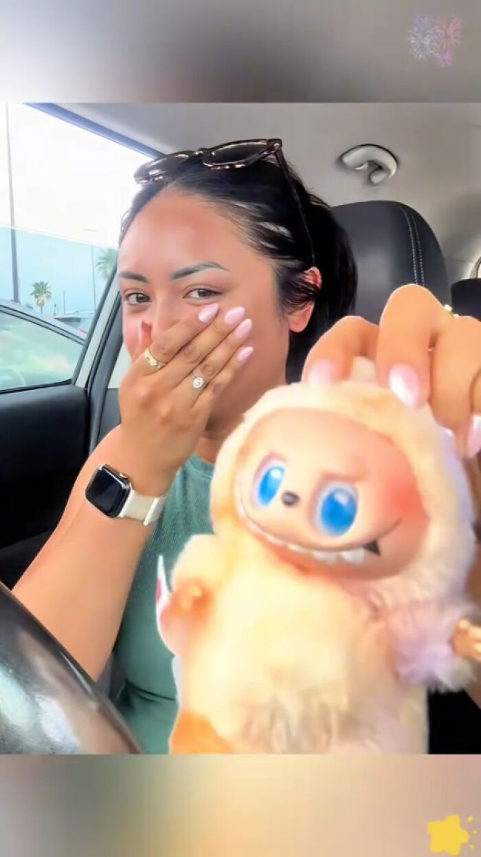 Woman in a car holding a creepy Labubus fake doll with large blue eyes and fluffy fur, reacting with surprise.