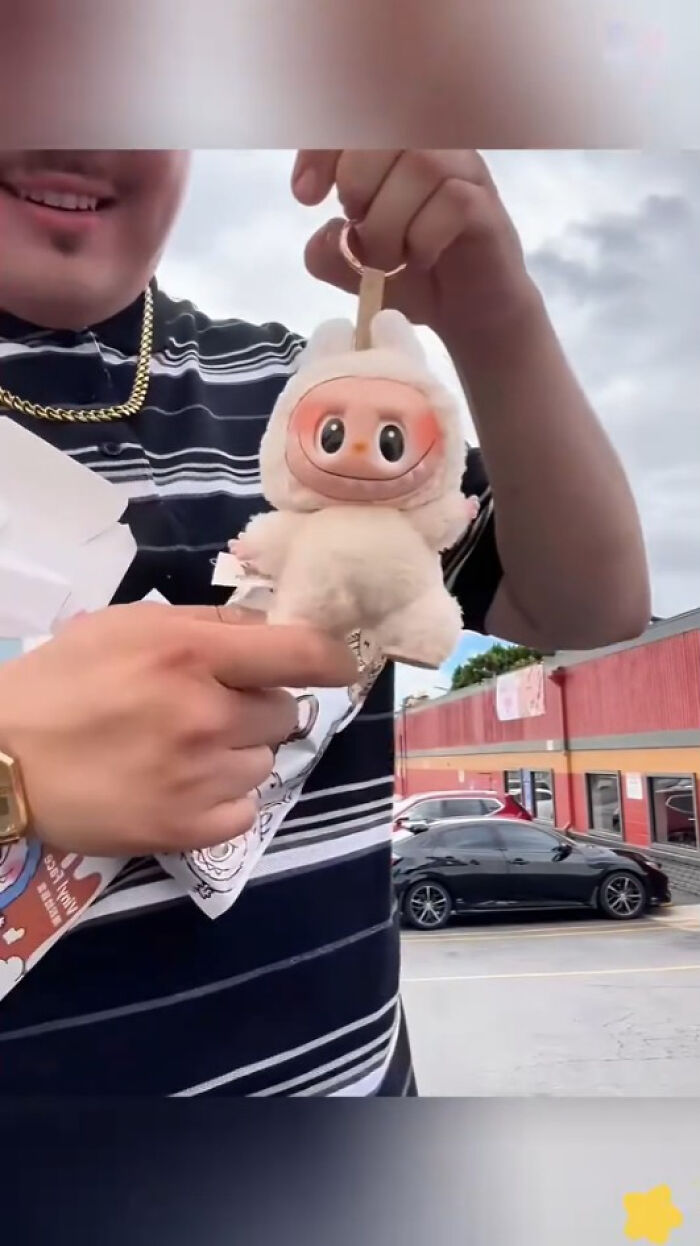 Person holding a creepy Labubus plush keychain with large eyes and a smiling face outdoors near a parking lot.