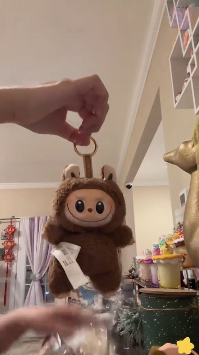 Hand holding Labubus keychain plush toy in a room with various colorful decorations and shelves in the background.