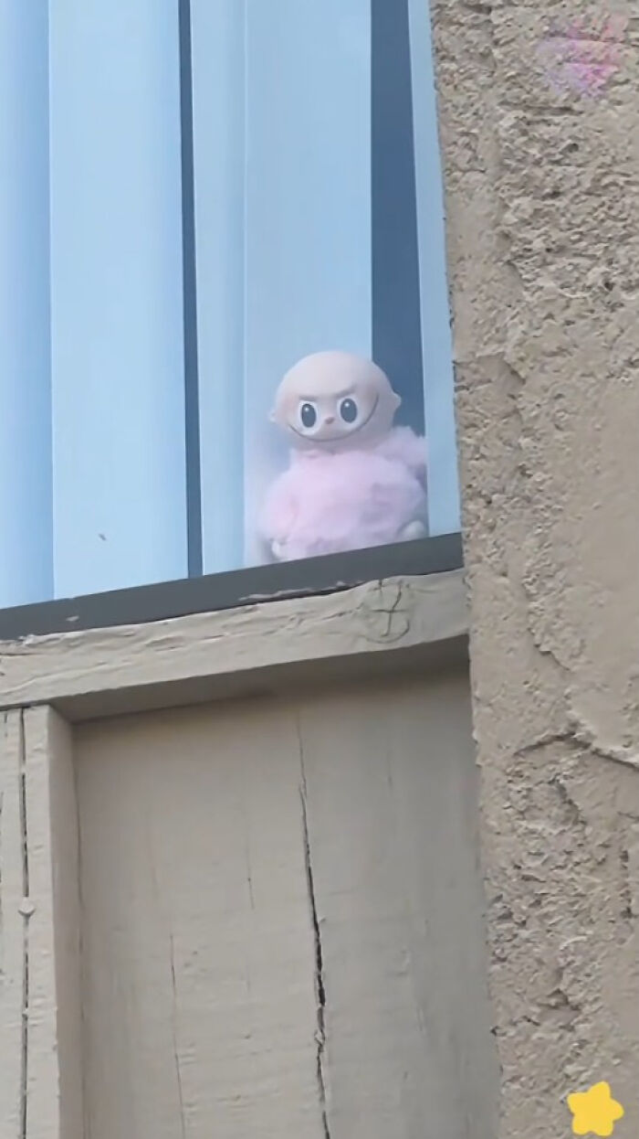 Creepy Labubus doll with large eyes and pink fuzzy body sitting behind vertical blinds in a window.