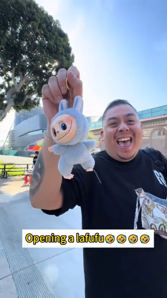 Man holding creepy Labubus fake plush toy outdoors, smiling widely on a sunny day with trees and buildings in background