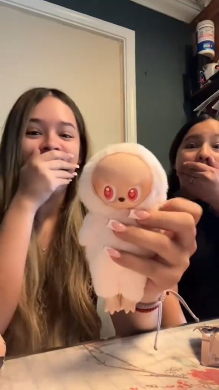 Two women react with shock holding a Labubus fake toy with red eyes in a dimly lit room.