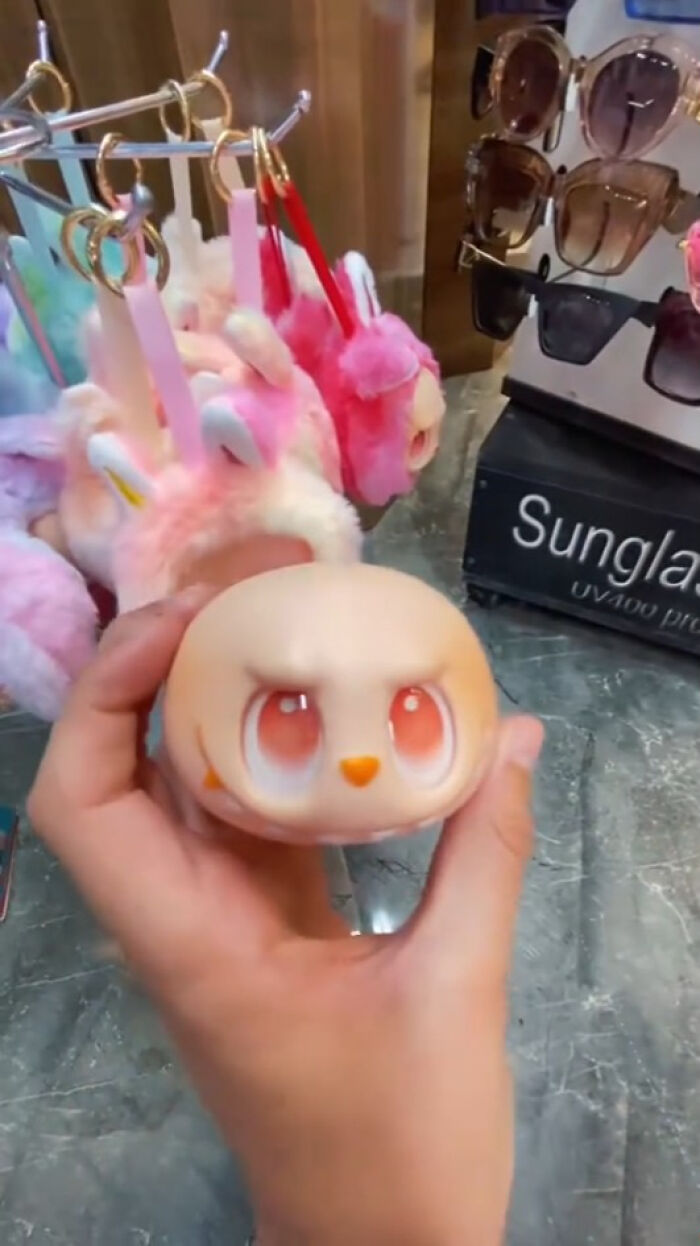 Hand holding a creepy Labubus fake toy with large eyes, displayed near colorful plush keychains and sunglasses stand.