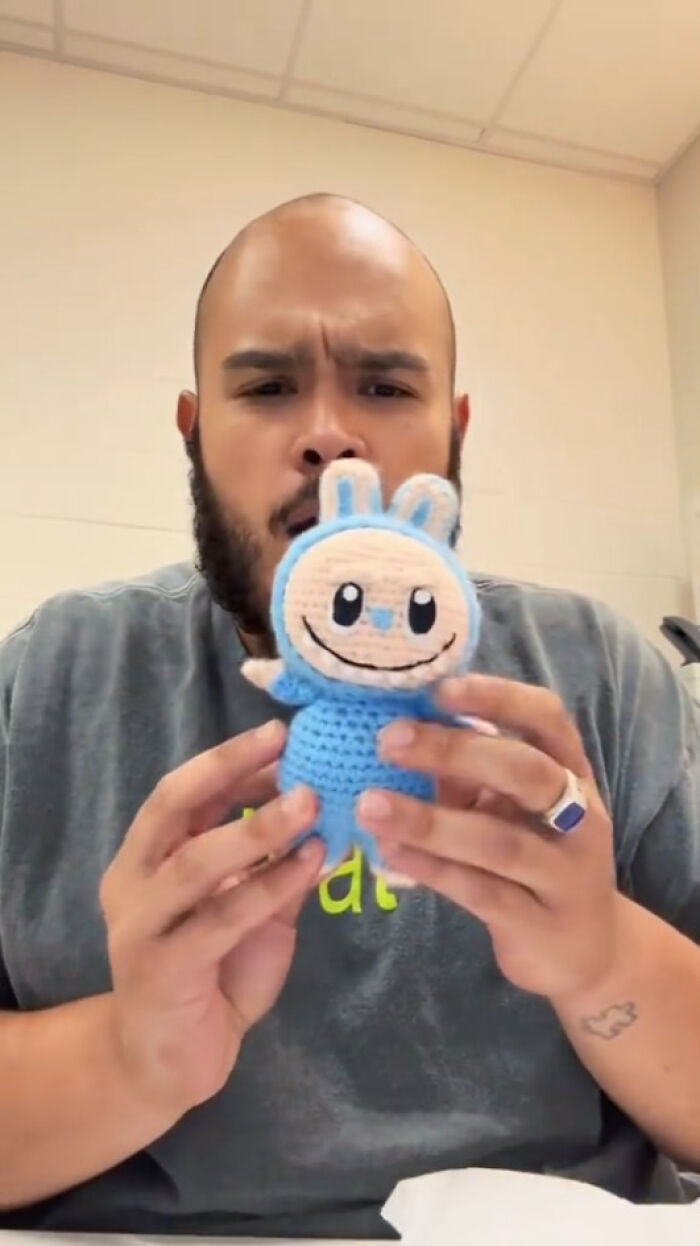 Man holding creepy Labubus fake doll with blue outfit and rabbit ears, showing handmade nightmare toy in indoor setting.