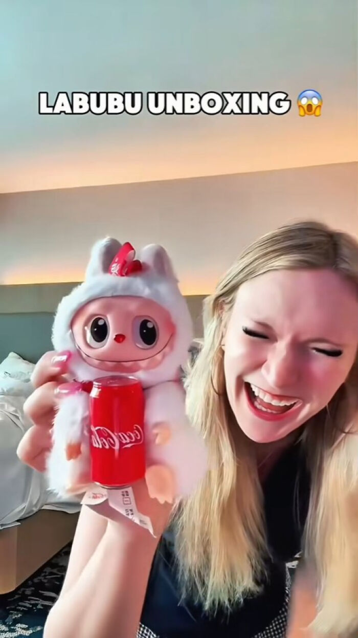 Woman unboxing a creepy Labubu fake doll holding a red can with playful expression in a modern room setting.