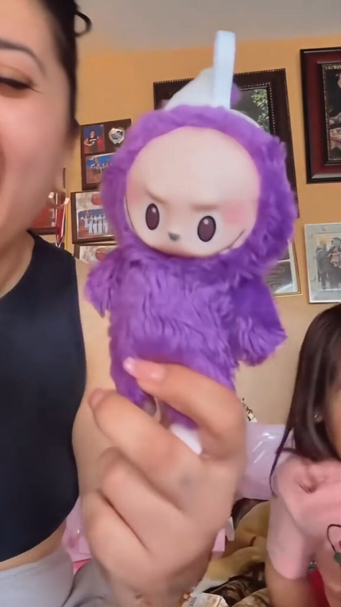 A person holding a creepy purple Labubus fake toy with an unsettling expression indoors.