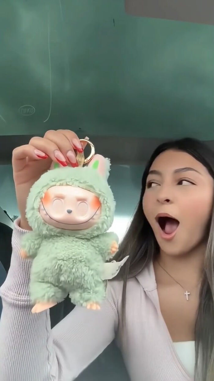 Young woman holding creepy Labubus plush keychain with exaggerated smile and green furry outfit inside a car.