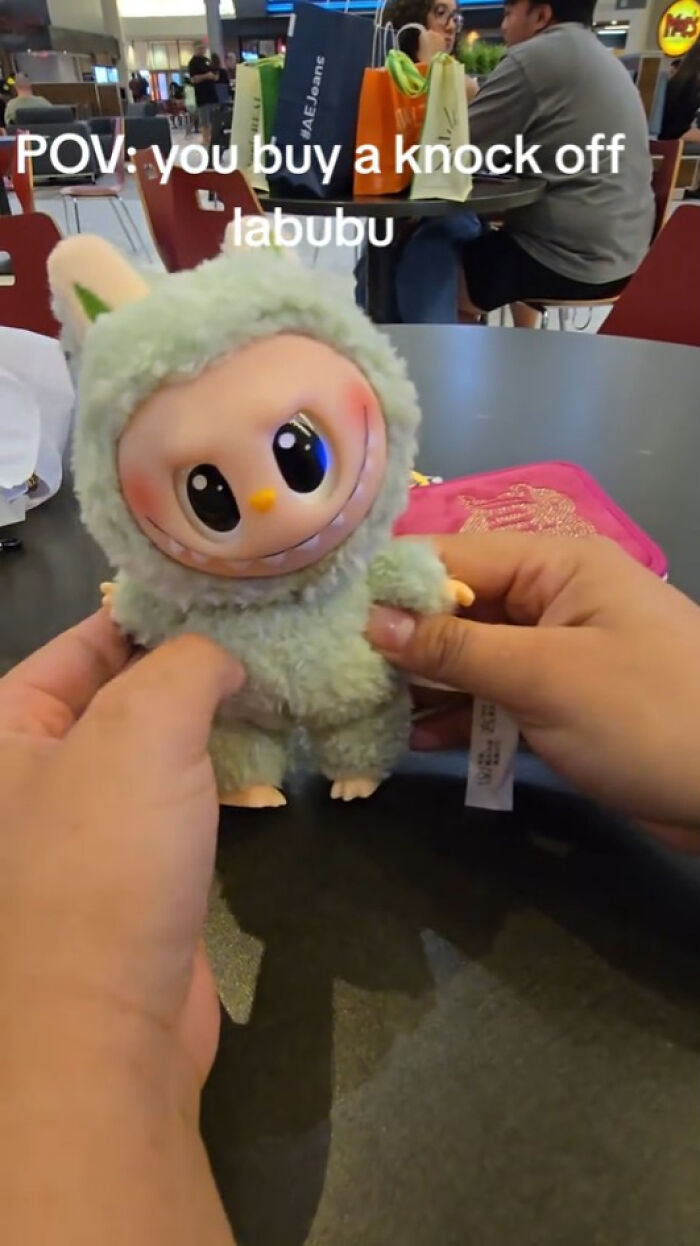 Hand holding a creepy green fuzzy Labubus knockoff doll with a wide smile and large dark eyes on a black table.