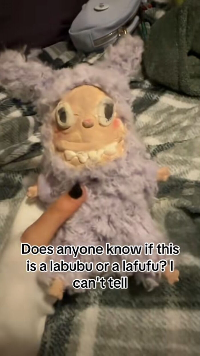 Hand holding a creepy Labubus plush with large eyes and sharp teeth, lying on a patterned blanket.