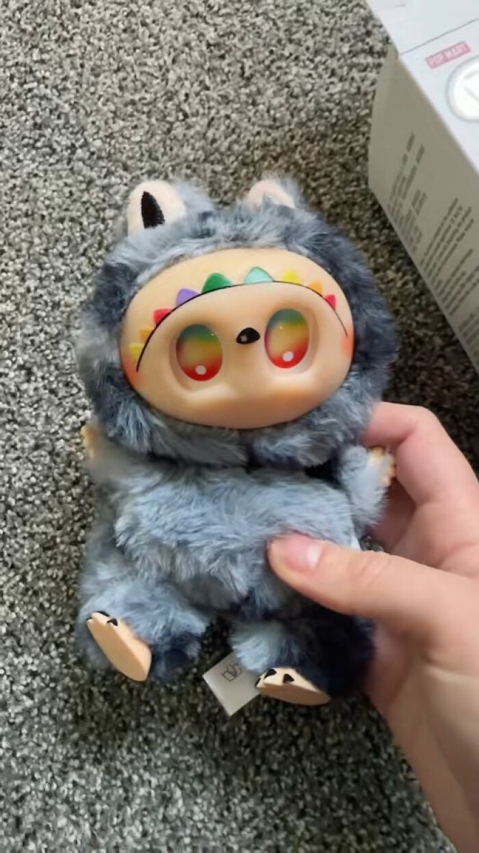 Hand holding a creepy Labubus fake doll with rainbow eyes and gray fur on a textured carpet background.