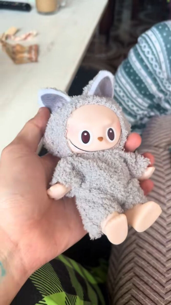 Hand holding a creepy Labubus doll in a fuzzy gray animal costume, highlighting fake nightmare-inducing toys.