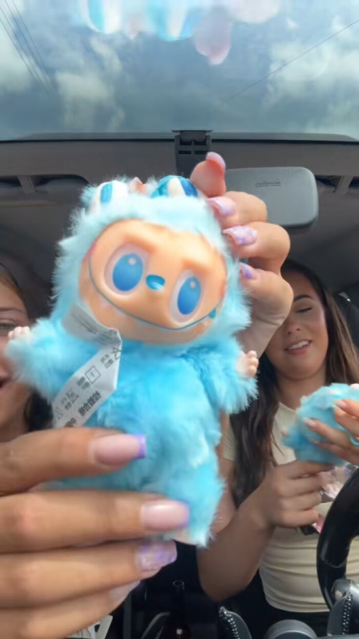 Two women in a car holding and showing creepy blue Labubus fake plush toys with exaggerated faces and features.