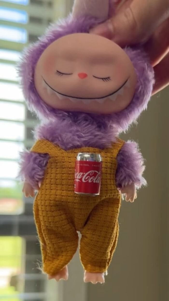 Creepy Labubus doll with purple fur and yellow outfit holding a small Coca-Cola can, evoking nightmare-like vibes.