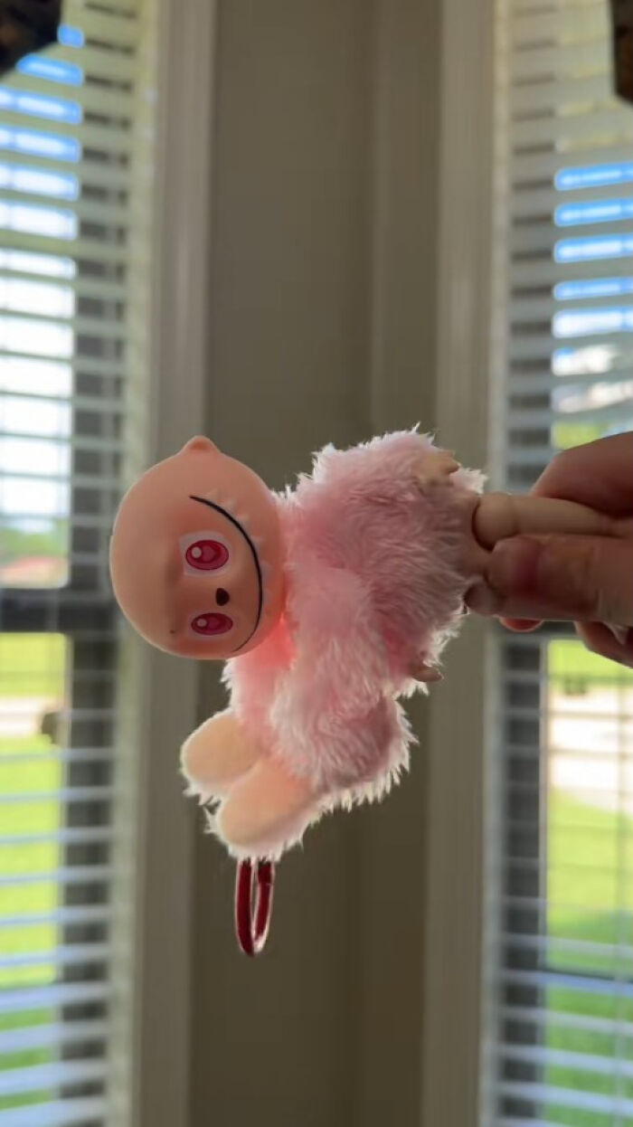 A creepy Labubus fake doll with pink fluffy body and unsettling face held indoors near window blinds.