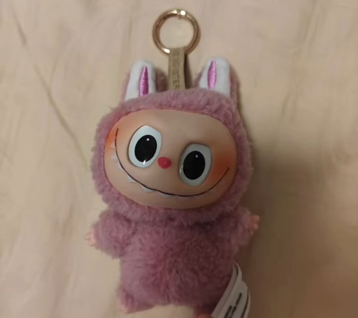 Plush toy keychain resembling a creepy Labubus fake with large eyes and a wide smile in pink fuzzy material.