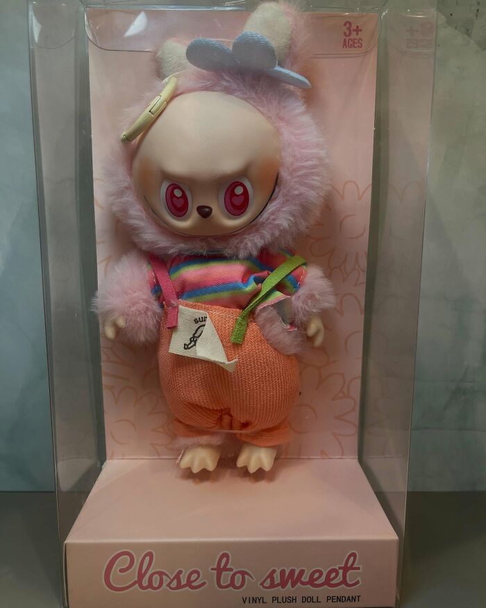 Creepy fake Labubus vinyl plush doll with pink fur, red eyes, and colorful outfit inside clear packaging.