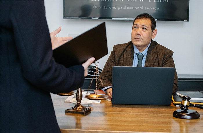 Lawyer in brown suit sitting at desk with laptop, scales of justice, and gavel, consulting a standing person in court context.