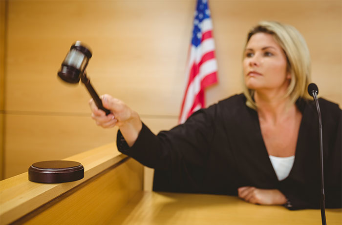 Female judge in courtroom raising gavel to make ruling with American flag in background showing court proceedings.