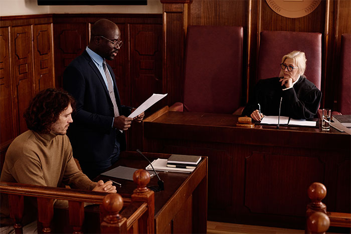 Lawyer reading documents in court with defendant seated nearby and judge listening attentively during trial session