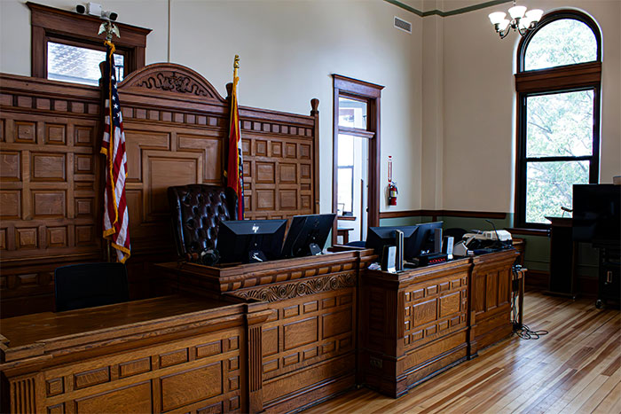 Wooden courtroom interior with judge’s bench, computer screens, flags, and large windows in a legal setting.