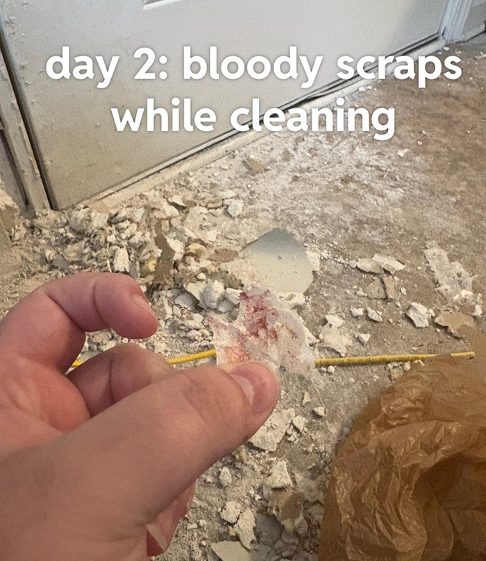 Hand holding bloody scraps while cleaning a dusty floor with debris and a brown bag nearby on day two.