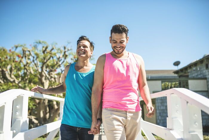 Two men wearing bright tank tops, smiling and holding hands while walking outdoors on a sunny day. - 9