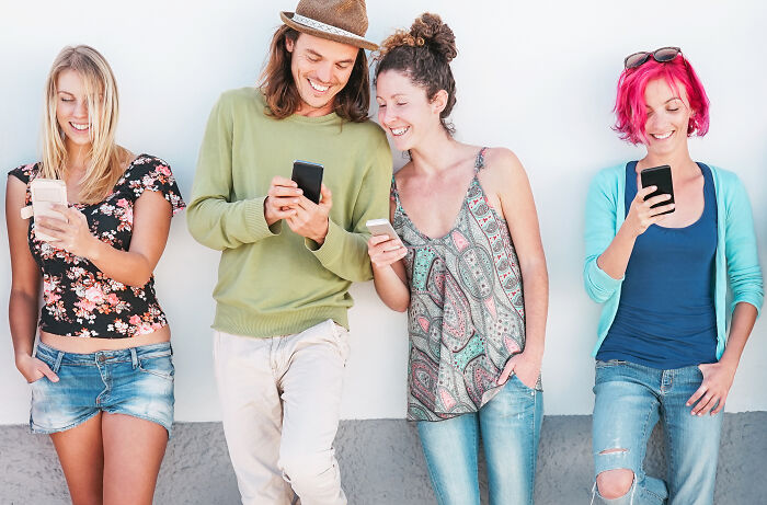 Group of Gen Z friends casually handing out compliments while smiling and using their smartphones outdoors against a white wall.